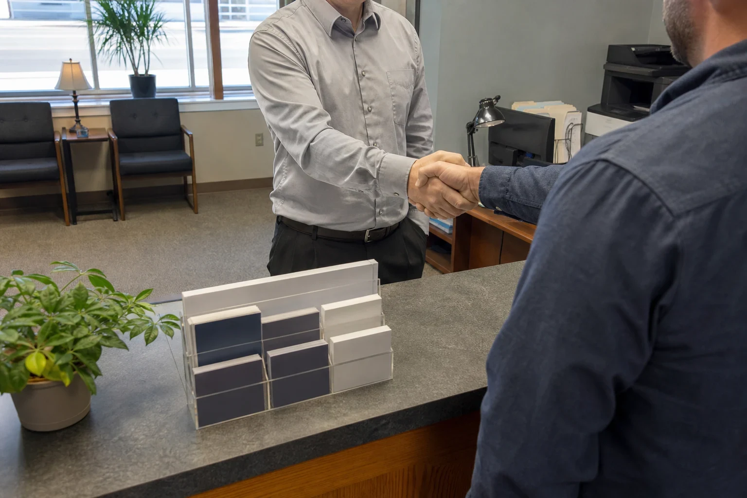 Agent in a light gray shirt shakes hands with a client across a reception counter in a carpeted office with guest chairs, a lamp and a front window