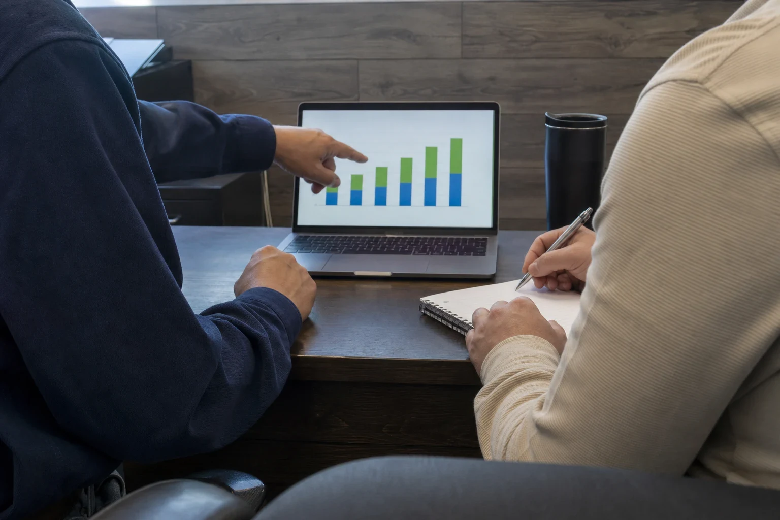 Two people at a dark wood office desk; one points at a bar chart on a laptop while the other takes notes with a pen in a spiral notebook